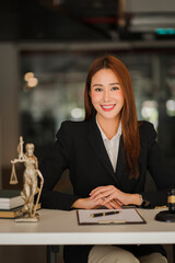 A beautiful Asian woman lawyer sits at her desk and looks at the camera taking notes of sample contracts with a hammer and the goddess of justice with a laptop at the Law Counselor Ideas Office.