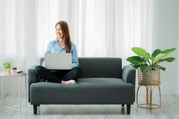 Fototapeta premium A happy Asian woman using a laptop computer sits on the sofa in the living room. work from home concept