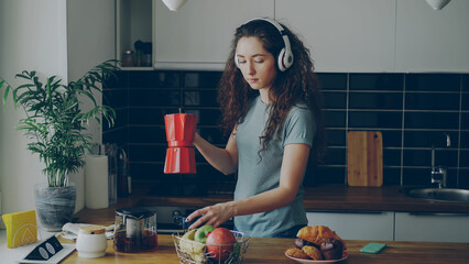 Curly beautiful caucasian woman in headphones dancing prepearing breakfast in kitchen at home