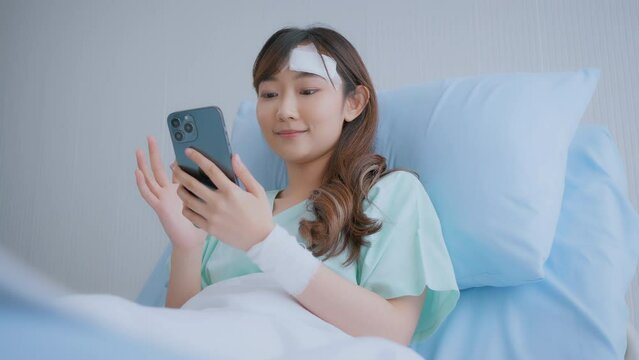 A young Asian woman had a head accident Currently using a smartphone and being admitted to the hospital. woman wearing a patient gown sitting in bed playing smartphone at the hospital