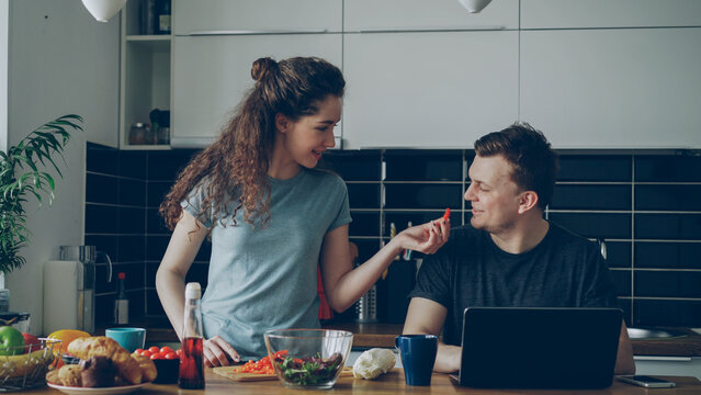 Cheerful couple at table woman cooking while gives hasband to try red pepper, smiling man sitting working on laptop