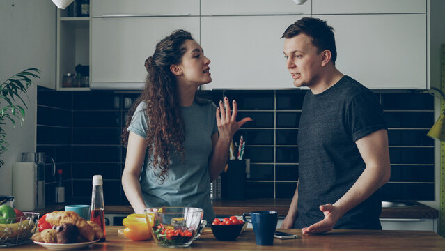 Pretty woman standing at table cutting salad while her boyfriend comes and they argueing speaking loud and nervous. Relations , difficulties concept - Powered by Adobe