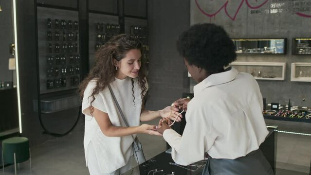Medium long slowmo of curly-haired Biracial woman standing by display in modern jewelry store, looking at necklace in hands of Black female consultant