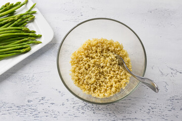Glass bowl with bulgur and a plate with asparagus on a light blue background. Cooking Delicious Homemade Vegan Asparagus Tabbouleh Salad