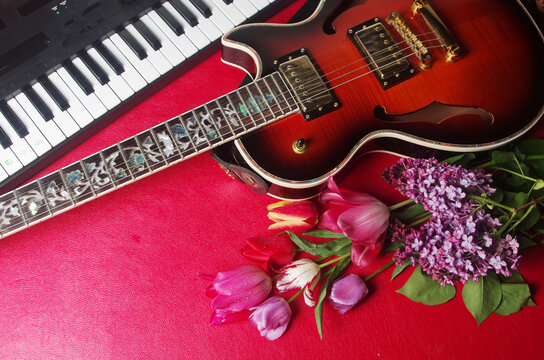 Electric guitar, violin, synthesizer keyboard and a bouquet of tulips and lilies of the valley on a red table. 
