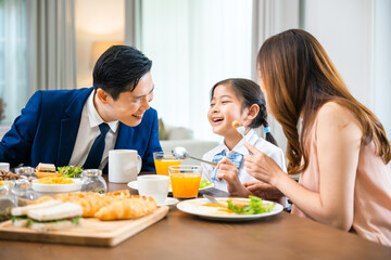 Asian family father, mother with children daughter eating breakfast food on dining table kitchen in mornings together at home before father left for work, happy couple family