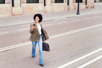 smiling business woman walking across the street checking the mobile phone with a briefcase in her...