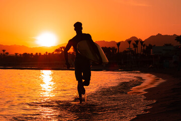 Silhouette man surfer in sunset light running down beach with surfboard near waters edge enjoying...