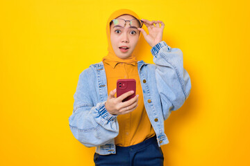Shocked young Asian woman in jeans jacket holding mobile phone, taking off glasses with opening mouth, reacting to great sale offers isolated over yellow background