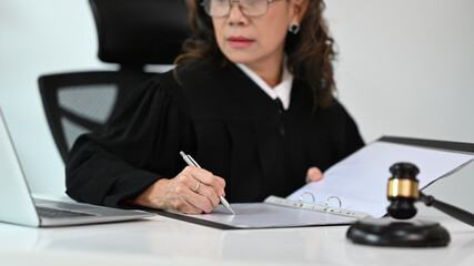Mature female judge, lawyer or attorney dressed in robe gown uniform sitting at table with gavel hammer mallet working with law document