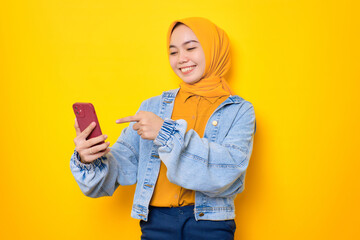 Cheerful young Asian woman in jeans jacket looking at mobile phone screen, raised hand with opening mouth, reacting to great news isolated over yellow background