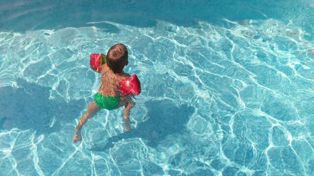A kid in oversleeves learns to swim in a children's pool with clean water, funny moving his legs on a summer day
