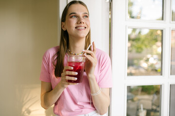 Cute teen girl holding a glass of juice and smiling at the camera. Smiling brunette in a pink t-shirt drinking lemonade posing and laughing