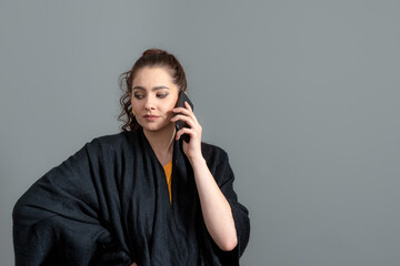 Fototapeta na wymiar curly-haired emotional young woman in a black cloak talk on mobile phone, isolated on gray