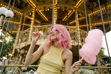 A cheerful girl in pink glasses eating cotton candy. Girl with pink hair posing with cotton candy. Carousel in the background. Rest in the amusement park