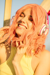 Close portrait of a girl listening to music in headphones. A happy girl with pink hair and a big smile. Amusement park in the background.