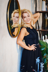 an attractive blonde in a black dress against the wall with a round mirror.