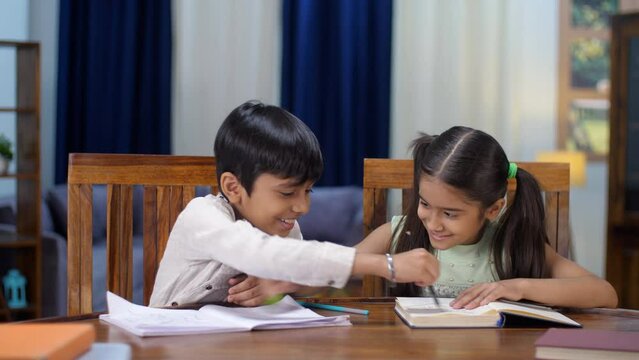 Young Indian brother-sister studying together while sitting at the dining table - knowledge and education. Cheerful siblings scribbling on each other's notebooks while doing their homework - a fun ...