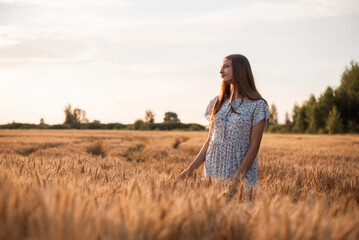 Naklejka na ściany i meble Beautiful dreamy girl in the field of rye at sunset. Portrait of a teenage girl admiring beauty of nature
