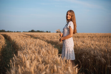 Side photo of a beautiful girl with blond hair holding spikelets of ripe rye looking to the camera. Portrait of a young girl walking in agricultural field on tractor traces