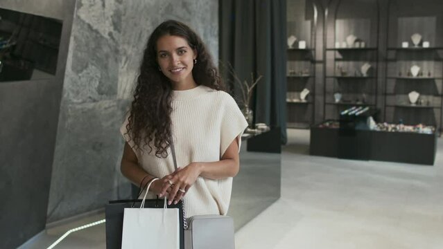 Medium long portrait of happy Biracial woman in white holding shopping bags, standing in luxury jewelry store, looking and smiling on camera