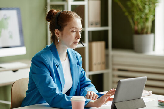 Young businesswoman in headset using digital tablet for online meeting with her colleagues at her workplace at office