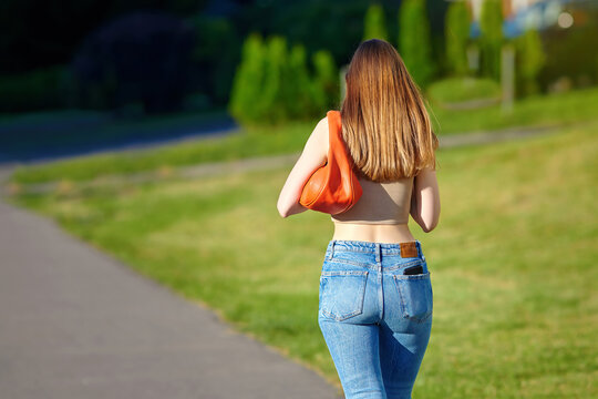 Stockfoto Young woman in stylish tight jeans, wearing crop top with handbag  walking outdoors in summer season, rear view. Woman wearing blue skinny  jeans and crop top shirts walking on city street.
