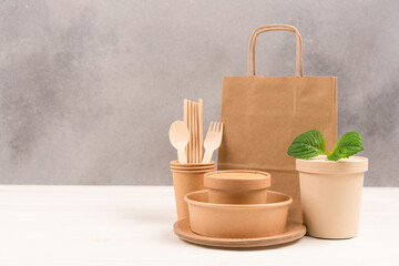 Paper utensils bundle - paper plates, food containers, bag, cups and wooden cutlery set against gray wall background with copy space. Sustainable food packaging. Eco tableware