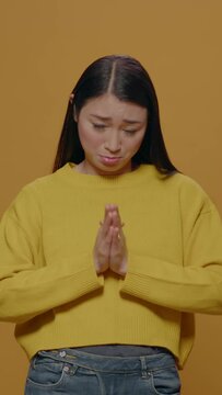 Vertical video: Asian woman doing praying gesture with hands and begging in front of camera, showing desire and expectation sign. Person imploring and asking with palms and body language in studio.