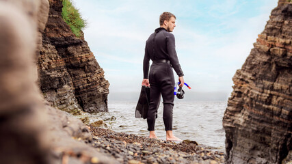a diver in a wetsuit stands on the seashore. Diver before diving into the ocean, in the hands of a mask and fins for diving