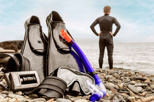 Flippers mask and cargo for scuba diving lie on the rocky seashore. In the background, a male diver stands with his back to the camera and looks towards the ocean.