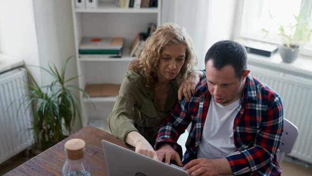Young man with Down syndrome sitting at desk in office and using laptop, his mother helping him.