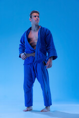 Portrait of young muscular man, martial art coach in blue kimono posing isolated over blue studio...