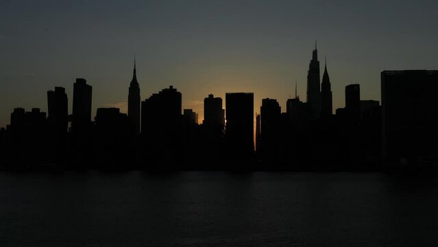 slow trucking shot of a silhouetted Manhattan skyline at sunset, just after the rare Manhattanhenge, a small sunbeam creeps from between the buildings