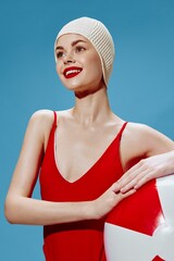 Inspired by the sea, a young woman in a retro red swimsuit smiling looks to the side holding a ball...