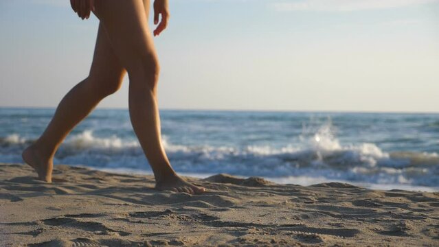 Close up of female feet walking at sea beach on sunny day with waves at background. Legs of young woman stepping at sand. Barefoot girl at seashore. Concept of summer vacation or holiday. Slow motion
