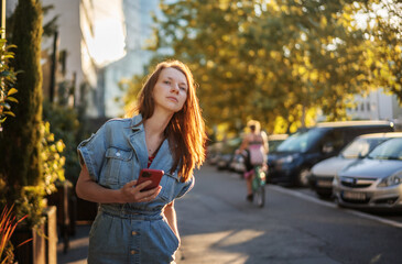 Young beautiful woman on a summer street in the city at sunset using smartphone