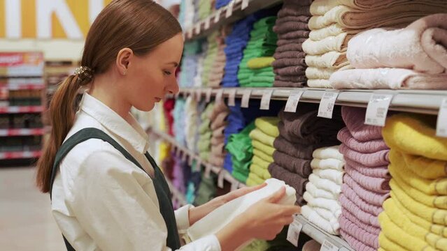 Young customer picks bright beige terry towel and checks material of item in household goods department. Red-haired woman enjoys going shopping in mall closeup