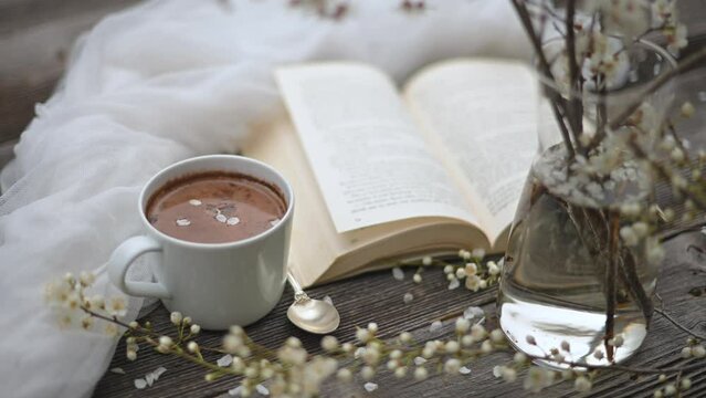 Cup of hot chocolate cacao, old book and spring cherry flowers on wooden background. The wind turns the page. Reading concept. UHD 4k video