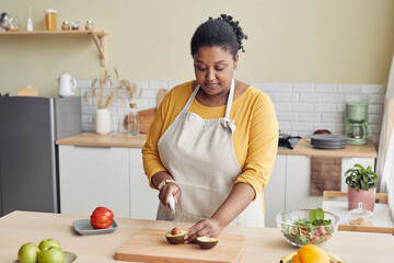 Portrait of young black woman cutting avocado while cooking healthy meal in kitchen, copy space