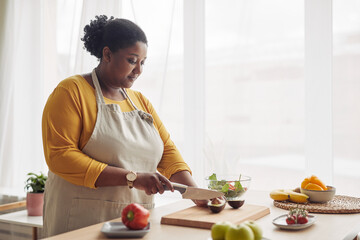 Portrait of young black woman cutting avocado while making salad in sunlit kitchen, copy space - Powered by Adobe