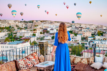 young woman in blue dress stands on terrace of hotel in Goreme Cappadocia and looks at hot air...