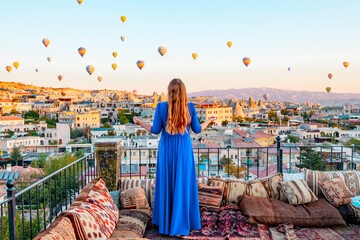 young woman in blue dress stands on terrace of hotel in Goreme Cappadocia and looks at hot air...