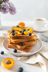 Tall stack of French toasts covered with blueberries, apricots and maple syrup on white background with lilac flowers. Breakfast menu. Bakery, restaurant. Copy space