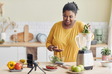 Portrait of smiling black woman using blender while making healthy meal in kitchen and filming...