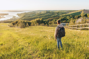 Fototapeta na wymiar Tourist with backpack standing on top of hill in grass field and enjoying beautiful landscape view. Rear view of teenage boy hiker resting in nature. Active lifestyle. Concept of local travel