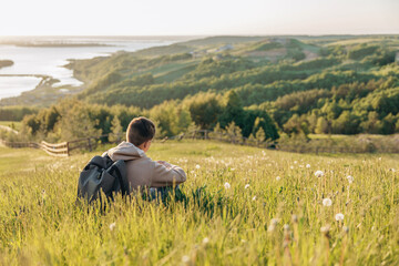 Fototapeta na wymiar Tourist with backpack sitting on top of hill in grass field and enjoying beautiful landscape view. Rear view of teenage boy hiker resting in nature. Active lifestyle. Concept of local travel