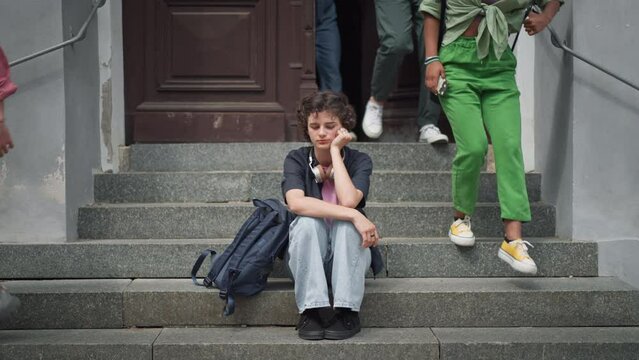 Sad teenager student sitting on stairs in front of school, listening to music. Bullying, discrimination or stress concept.