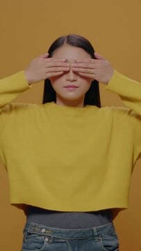 Vertical video: Asian woman covering mouth, eyes and ears in studio, doing three wise monkeys symbolic gesture, not listening to noise and not speaking. Young adult gesticulating serious expression