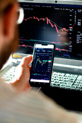 View from the back of man holding mobile phone in hand with financial graph monitoring stock. Online trading crypto currency. Working online from home. Data visualising. Selective focus